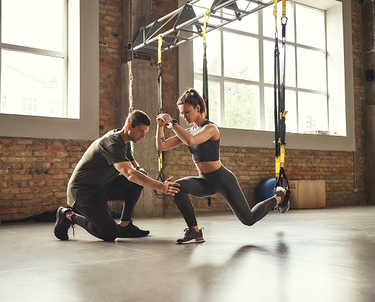 Entraînement personnel TRX : perfectionnez vos fentes. Une athlète fait des fentes bulgares avec des sangles TRX, assistée par son coach personnel dans une salle de sport style loft.