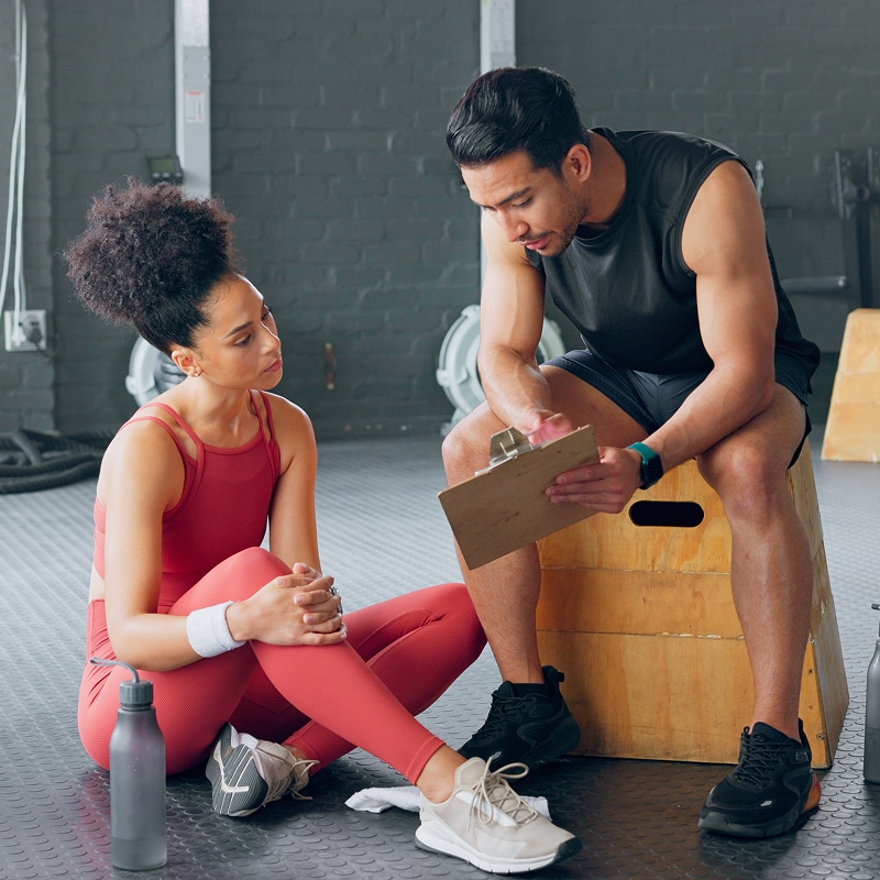 Consultation fitness intense : l'entraîneur personnel, assis, examine le programme sur un presse-papiers tandis que l'athlète écoute au sol.