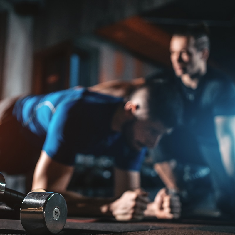 Athlète en T-shirt bleu fait la planche (gainage) lors d'un entraînement intense. Un coach le supervise, près d'un haltère « 5 ».