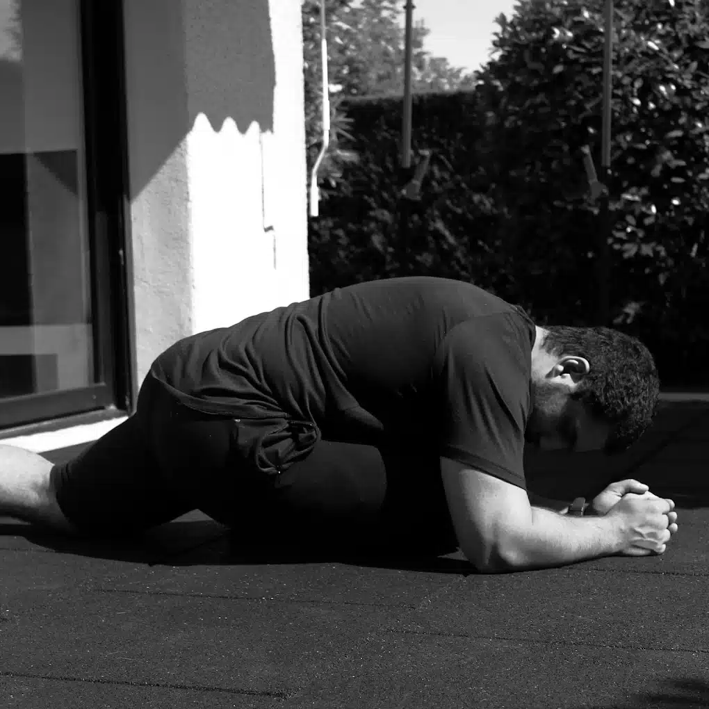 Étirement profond des hanches : Améliorez votre souplesse Entraînement : homme effectuant un étirement profond de la hanche (pigeon pose) sur un sol en caoutchouc noir, sous un soleil intense.