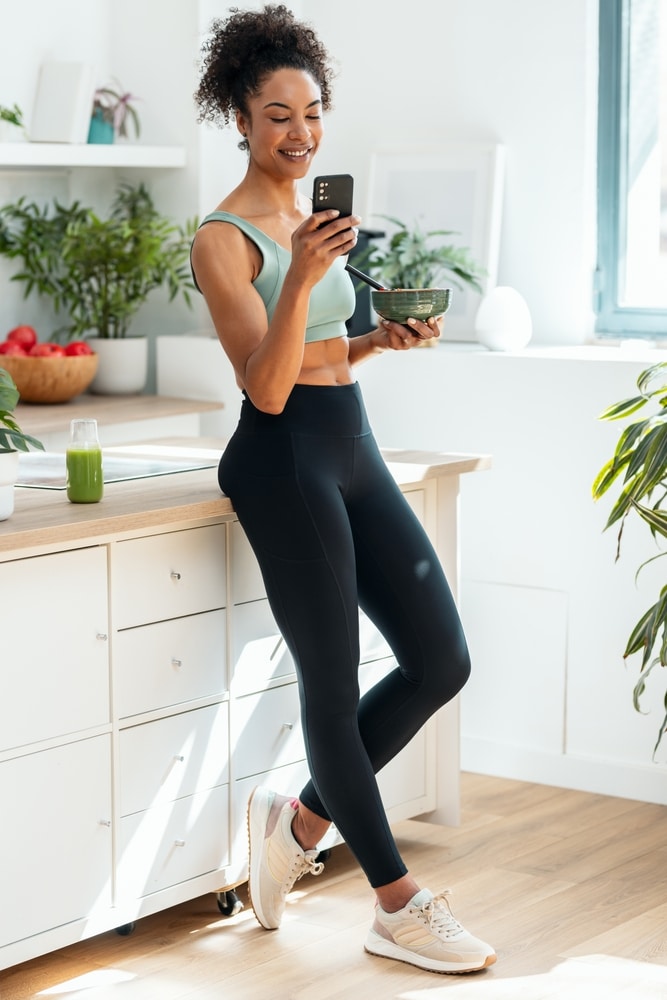 Fitness matinal : petit déjeuner healthy en legging. Femme fitness souriante, en brassière et legging noir, mange son petit déjeuner protéiné dans une cuisine lumineuse et regarde son téléphone.